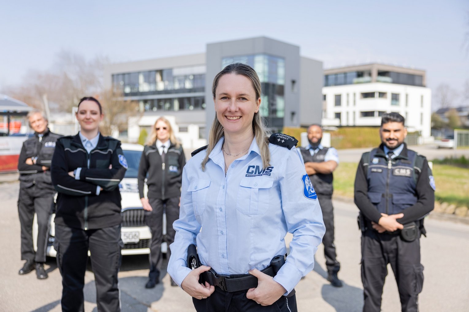 Mitarbeiterin des CMS Sicherheitsdienstes in Uniform steht vor Team und Einsatzfahrzeug vor modernem Gebäude